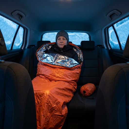 Kvinna sitter i baksätet på en insnöad bil med en orange nödsovsäck uppdragen till bröstet