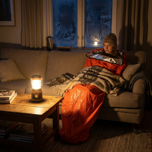 Tjej i soffan under strömavbrott med orange nödsovsäck och en ullfilt ovanpå, upplyst av en lykta