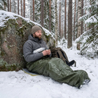 Man i grön nödsovsäck sitter på ett underlag i snön mot ett stort block i tallskogen med en varm kopp