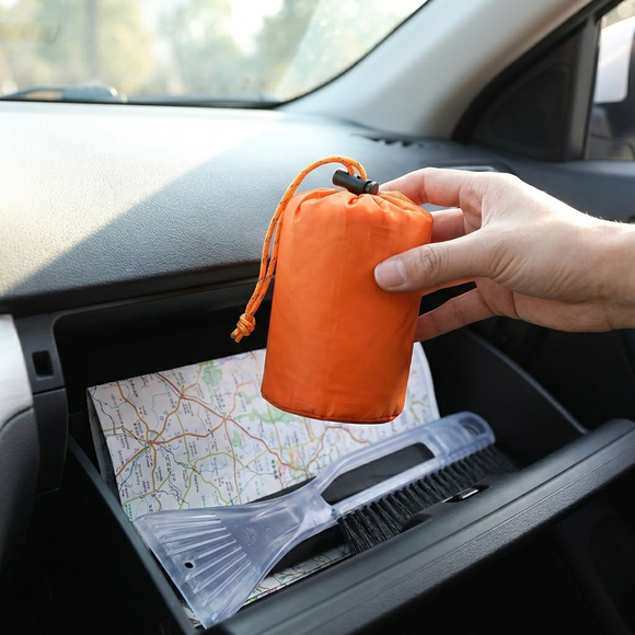 Hand lägger nödsovsäckens lilla orange fodral i bilens handskfack bredvid en karta och en isskrapa