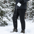 Vatten hälls från en emaljmugg på svarta vinterbyxor och pärlar sig och rinner av tyget i snön