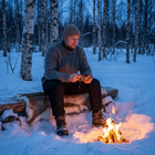 Man i svarta vinterbyxor sitter på en renfäll vid en lägereld i snöig björkskog i skymning