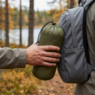 Hand håller den packade gröna filten i sin påse bredvid en liten grå dagsryggsäck i höstskog