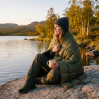 Kvinna sitter på en klippa vid en fjällsjö med grön pufferfilt över axlarna och en emaljmugg i handen