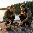 Par steker fisk i stekpannan på ett litet stormkök på en klippa vid sjön i solnedgången